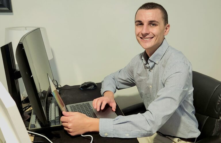 A picture of Bradley Hlibok sittng at a computer. He is a white male with short hair smiling to the camera.
